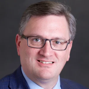 Professional portrait of a middle-aged man wearing glasses, a blue checkered blazer, and a light shirt, smiling warmly.